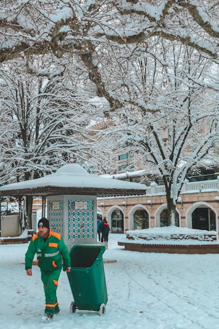 A waste collection worker dressed in a green and orange high-visibility uniform is pushing a wheeled green rubbish bin along a snow-covered pavement in an outdoor setting. Behind him, there is a small covered structure with a decorative tiled wall, and a few pedestrians walking in the background. The scene is framed by snow-laden trees with bare branches overhead, creating a winter landscape. The environment appears to be a public space, possibly a park or a town square, with surrounding buildings partially visible. The snow blankets the ground and the tops of nearby benches and planters, highlighting the cold weather. The presence of the waste worker and the rubbish bin suggests routine waste collection or disposal activity, aligning with private waste management services sometimes involving on-site clearing, especially during inclement weather. The overall scene emphasizes outdoor rubbish collection in winter conditions, characteristic of independent waste handling services that operate beyond regular municipal refuse collection.