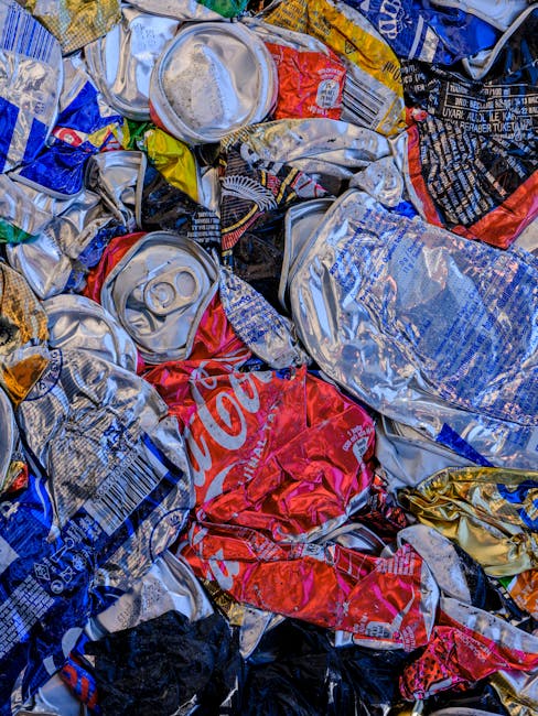 A waste collection worker wearing a high-visibility yellow vest with reflective stripes and dark trousers is operating the controls of a large rubbish collection truck, which is parked on a city street during evening hours. The truck's body is painted white with red and orange diagonal reflective safety stripes along the rear, and features blue and white branding on the side. The worker is focused on managing the loading process, with various bulging bags and loose waste visible in the truck's open hopper area. The environment includes a building façade with windows illuminated from within, casting a warm glow, contrasting with the darker, muted tones of the street scene. This scene exemplifies an alternative waste handling method involving private refuse collection rather than municipal rubbish disposal, aligning with the theme of avoiding hidden rubbish removal charges in Tooting Market jobs and showcasing a typical rubbish removal operation.