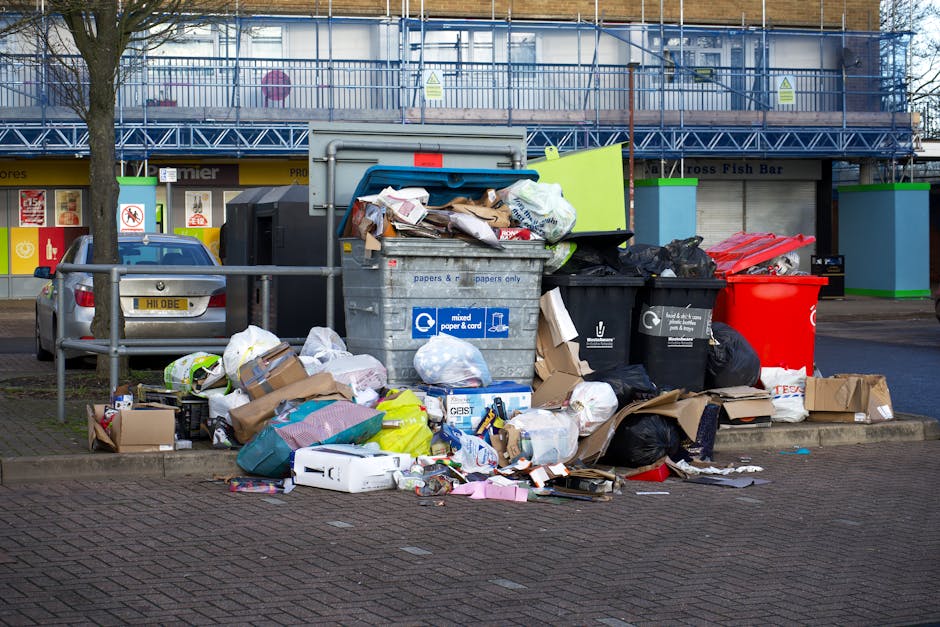 An outdoor scene in a parking area shows several overflowing trash bins and an assortment of garbage bags, cardboard boxes, and discarded packaging piled on the pavement. A large grey mixed paper and cardboard recycling bin with a blue lid and handle is central, with paper and cardboard spilling over its top and onto the ground. To its right, there are black rubbish bags, a red bin, and more flattened boxes, some with visible branding and labels. In the background, a blue and white construction scaffold surrounds a building, with a row of small shops featuring signage, including a fish bar, visible behind a low metal railing. A parked silver van is positioned to the left of the bins, with part of a tree and its branches extending into the frame. The overall scene highlights an unmanaged waste area potentially related to private rubbish collection services, with some rubbish scattered nearby, and the environment suggesting an urban setting.