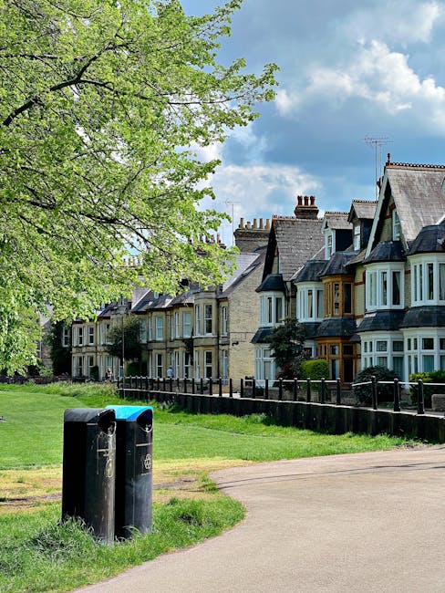 A row of traditional terraced houses with bay windows and brick facades line a quiet residential street. In the foreground, a large leafy tree with fresh green foliage extends its branches over a well-maintained lawn area. Two black public litter bins, made of plastic or metal with slightly rounded tops, are positioned on a paved pathway that runs parallel to the houses. The bins are slightly weathered, showing minor surface marks. The sky above is partly cloudy with patches of blue and soft white clouds, suggesting a clear, pleasant day. A wooden fence separates the grassy area from the pavement and houses, adding a boundary to the front gardens. The scene reflects a typical suburban neighborhood where residents or local authorities might undertake independent waste collection or rubbish disposal, supported by nearby rubbish removal services like Rubbish Removal Tooting. Overall, the image depicts a clean, orderly environment suitable for private waste handling, emphasizing community-maintained green spaces and urban living aesthetics.