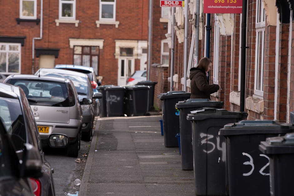 A row of black wheelie bins with closed lids lined up along a pavement next to a brick residential building, with some graffiti on the bins. A woman dressed in a brown coat is seen standing near the bins, possibly disposing of rubbish. Several parked cars, including small hatchbacks in silver and other dark colours, are parked along the street, which is narrow and lined with similar brick terraced houses in the background. The environment appears to be an urban residential area with a typical street scene, and the presence of multiple refuse bins suggests ongoing rubbish collection or waste removal activities as part of local or private waste management services. The lighting indicates natural daylight, with a neutral and professional tone suitable for a waste management context.