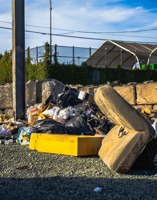 A pile of mixed rubbish including black plastic bags, a yellow plastic container, and a discarded beige upholstered armchair with visible fabric texture, all accumulated on a gravel surface outdoors. The background features a low stone wall, some green bushes, and a partly cloudy blue sky with a utility pole and overhead power lines. The scene suggests an instance of unauthorized waste dumping near a sports or event venue, with the rubbish left outside for collection by private waste removal services such as Rubbish Removal Tooting. The image emphasizes the need for professional rubbish clearance and highlights the importance of proper disposal methods for bulky and assorted waste materials.
