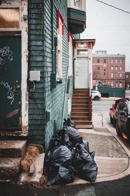 A row of black wheelie bins with closed lids lined up along a pavement next to a brick residential building, with some graffiti on the bins. A woman dressed in a brown coat is seen standing near the bins, possibly disposing of rubbish. Several parked cars, including small hatchbacks in silver and other dark colours, are parked along the street, which is narrow and lined with similar brick terraced houses in the background. The environment appears to be an urban residential area with a typical street scene, and the presence of multiple refuse bins suggests ongoing rubbish collection or waste removal activities as part of local or private waste management services. The lighting indicates natural daylight, with a neutral and professional tone suitable for a waste management context.